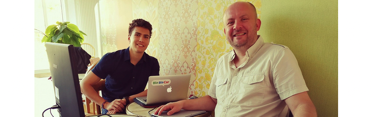 Two men sitting at a desk with laptops, engaged in work